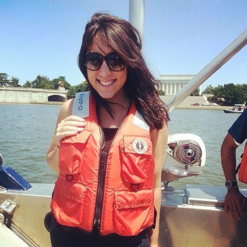The image shows a woman wearing sunglasses and an orange life vest on a boat. She is smiling and appears to be enjoying herself. In the background, there is a body of water, a bridge, and a building. The sky is blue and the weather seems pleasant.